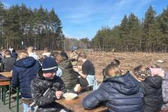 Uczestnicy-akcji-sadzenia-lasu-podczas-wspolnego-posilku-na-lonie-natury.-Dzieci-wraz-z-opiekunami-siedza-przy-lawkach-w-tle-widoczny-jest-wysoki-las